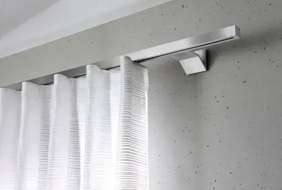 Detailaufnahme einer eckigen, silbernen Innenlauf-Vorhangstange an einer Sichtbetonwand mit weißem Wellenvorhang.