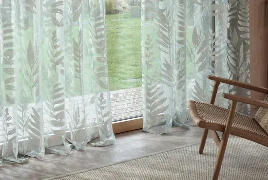 Leichte, weiße Gardinen mit dezentem Farnmuster vor einem Fenster mit Blick in den Garten.