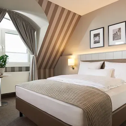 Blick in ein helles Schlafzimmer mit Dachschräge, gestreifter Tapete und farblich abgestimmten grauen Vorhängen.