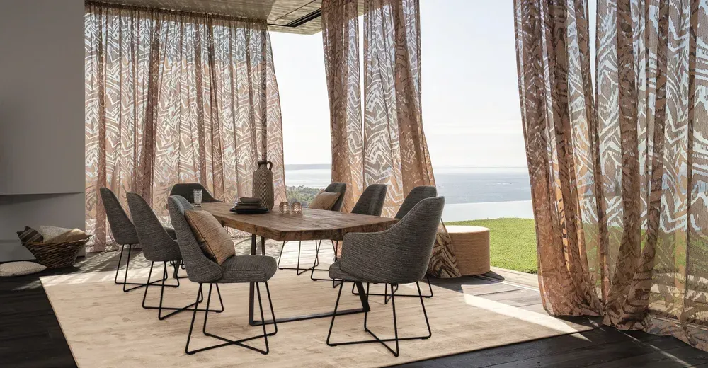 Eine Terrasse mit modernen Flechtsesseln und einem großen Esstisch, geschützt durch leichte, dekorative Outdoor-Vorhänge.