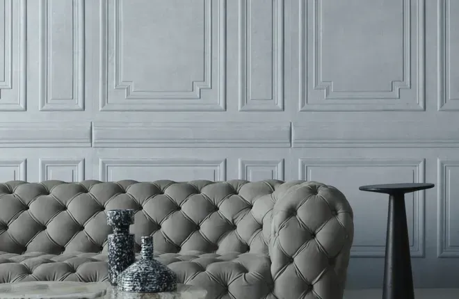 Eine hellgraue Wand in edler Kassetten-Optik (Stuckleisten), davor ein klassisch kapitoniertes Sofa in der gleichen Farbe.
