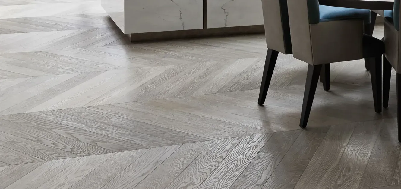 Detailaufnahme eines hochwertigen Holzfußbodens im Fischgrätenmuster mit den Beinen eines modernen Tisches und Stuhls.