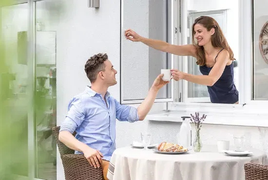 Ein Paar interagiert an einem offenen Fenster mit Fliegengitter; der Mann sitzt innen am Tisch, die Frau steht draußen.