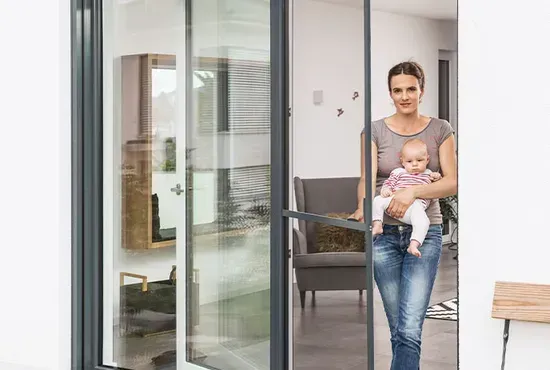 Eine Frau mit einem Baby auf dem Arm geht durch eine geöffnete Fliegengitter-Tür auf die Terrasse.