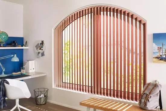 Ein Arbeitszimmer mit einem Rundbogenfenster, das mit vertikalen Holz- oder Stofflamellen in warmem Braunton beschattet wird.