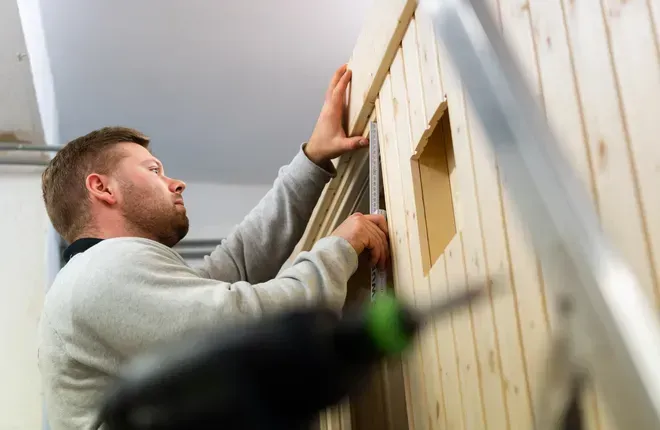 Ein Fachmann montiert präzise Holzpaneele an einer Wand, im Vordergrund ist unscharf ein Akkuschrauber zu sehen.