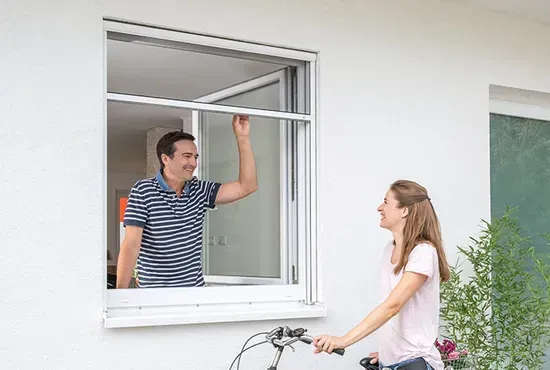 Ein Mann bedient ein vertikales Fliegengitter-Rollo an einem Fenster, während eine Frau draußen zuschaut.