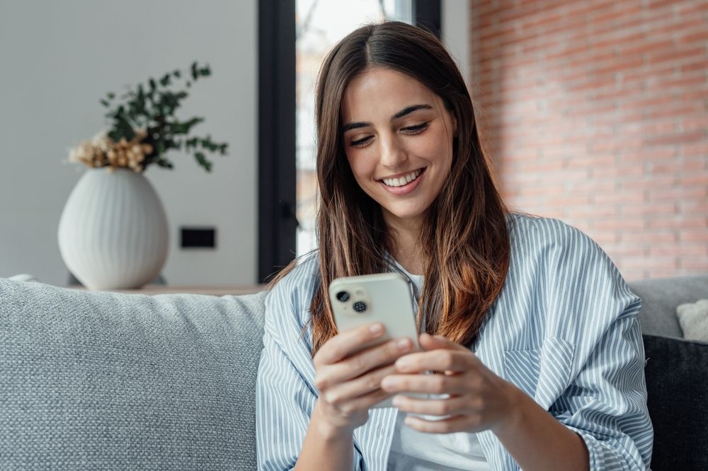 Una persona sonriente con cabello castaño largo mira fijamente un teléfono inteligente de color claro mientras está sentada en un sofá gris.