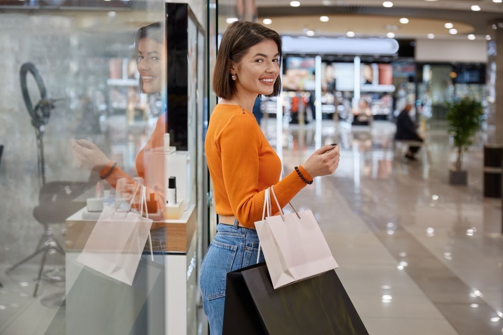 Una compradora sonriente, vestida con una camisa naranja de manga larga, sostiene bolsas de compra blancas y negras mientras está de pie en un centro comercial.