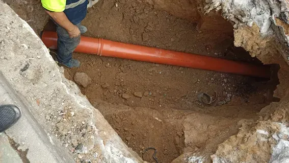 Un hombre está parado en un agujero en el suelo al lado de una tubería naranja.