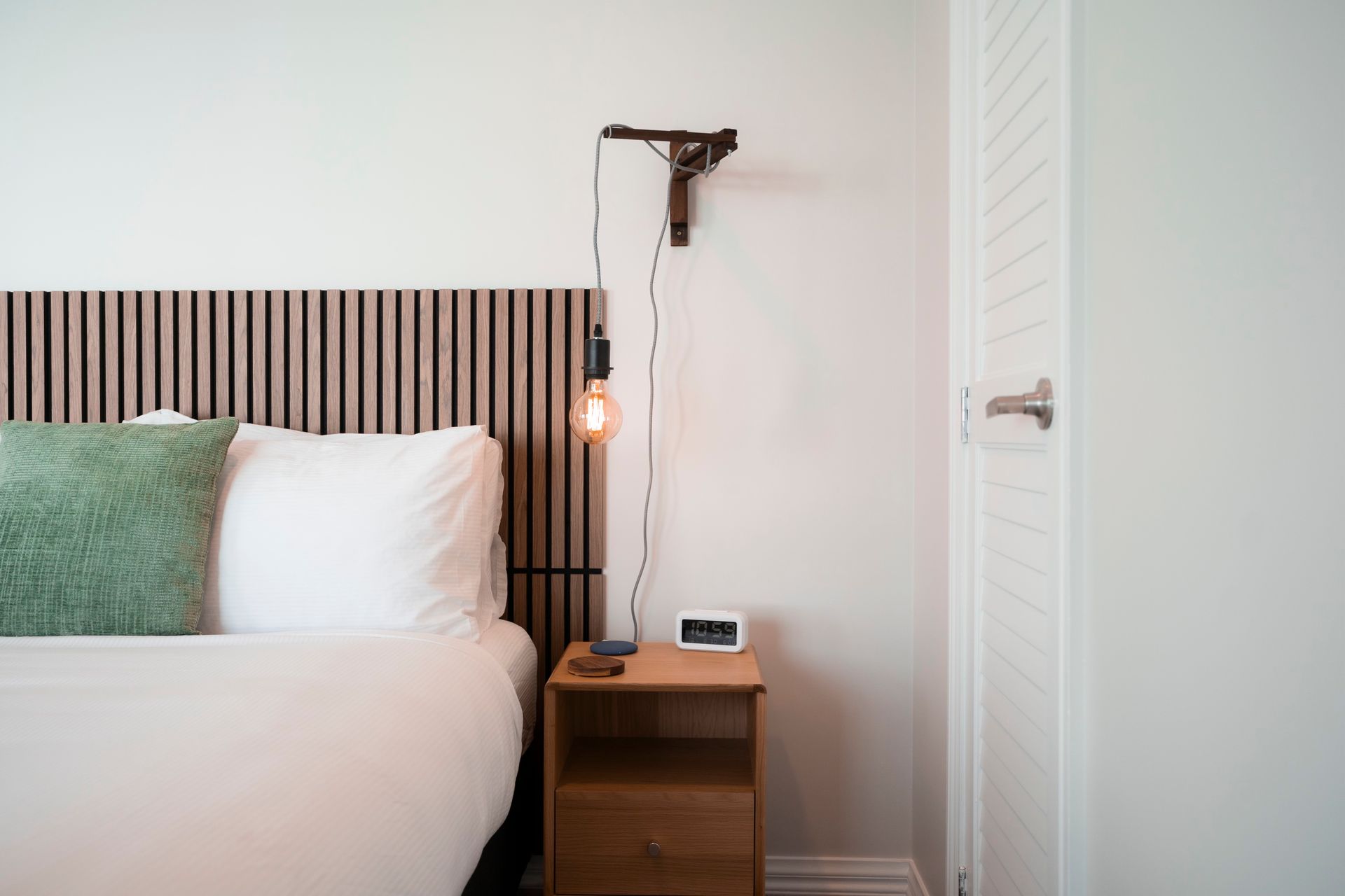 Bedroom with bed, nightstand, and wall-mounted light. Green pillow, wooden headboard, and white door.