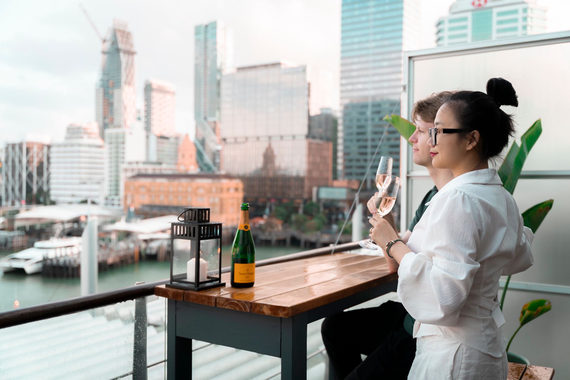 Couple toasting with champagne on a balcony, overlooking a cityscape with a harbour.