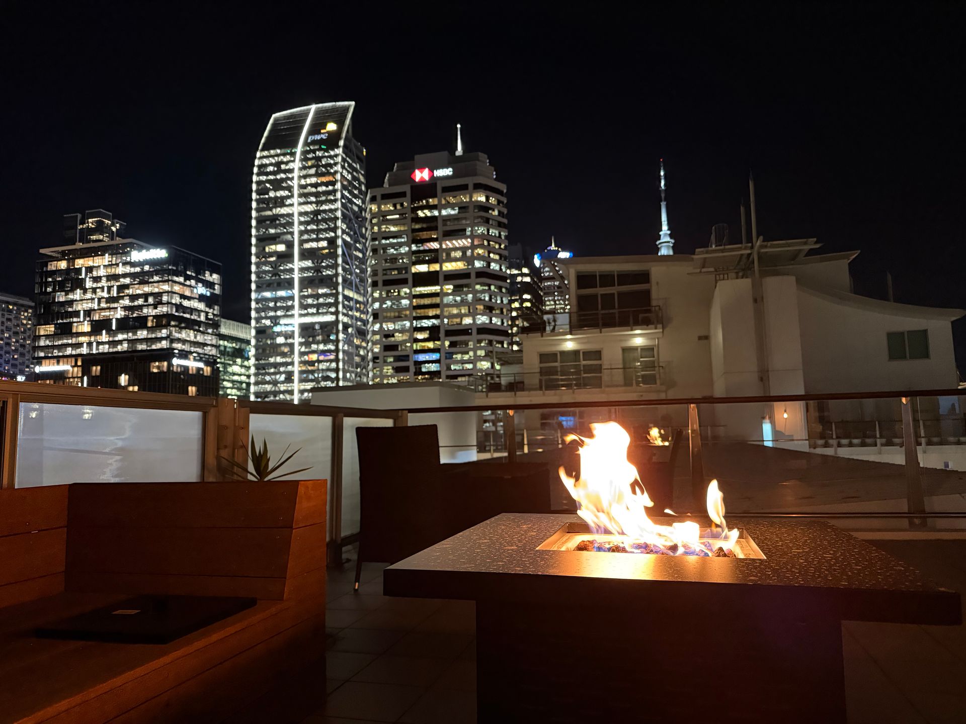 Rooftop fire pit with city skyline at night, featuring lit skyscrapers and seating.