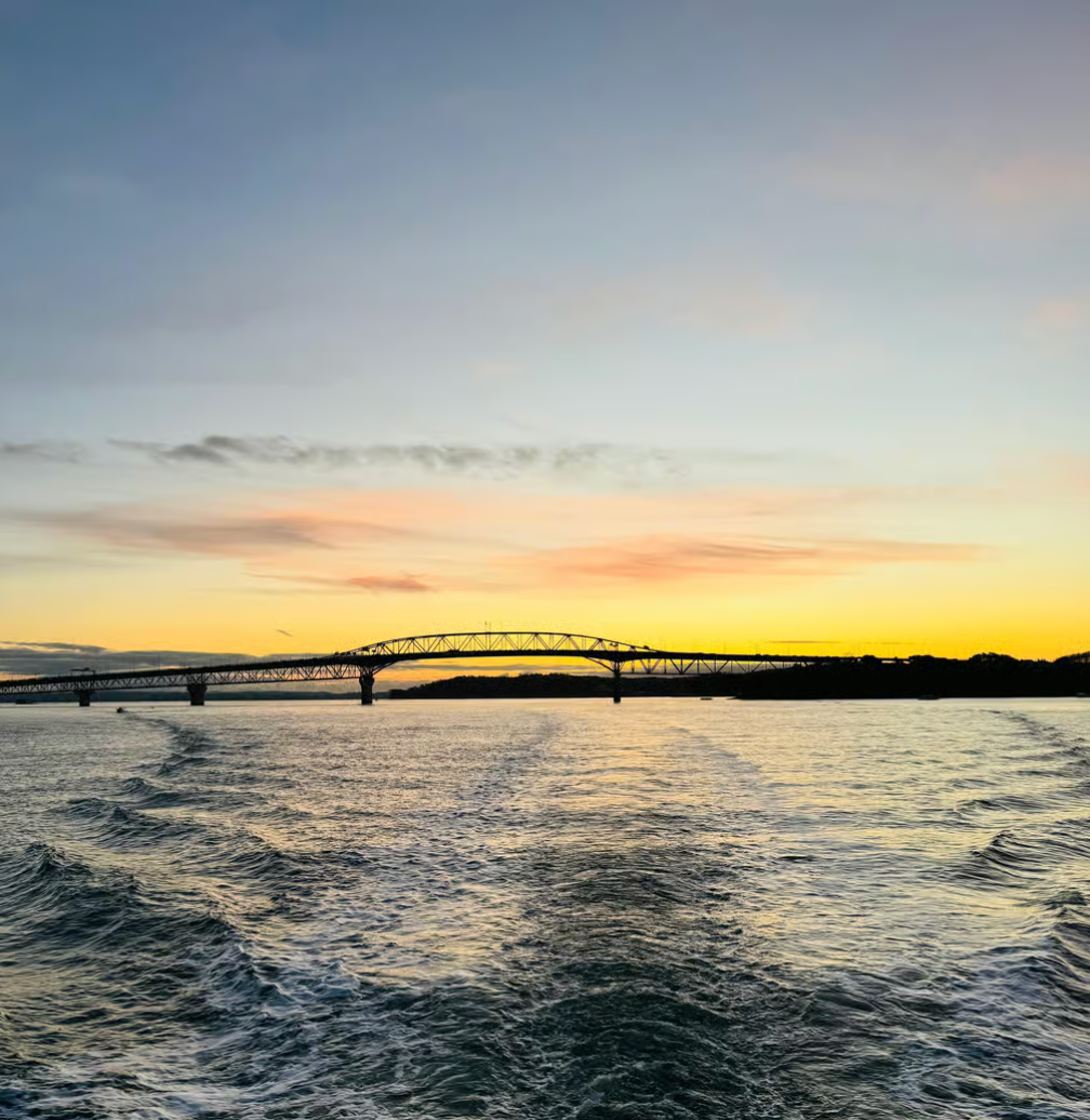 A bridge stretches across calm water at sunset, with a wake trailing behind the viewer towards the horizon.