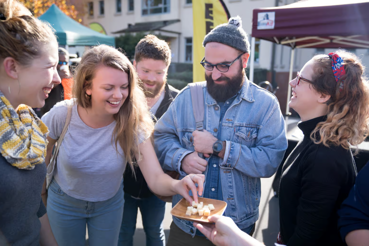 A group of people laughing together outdoors at a market event, while one person reaches for a snack on a wooden tray.
