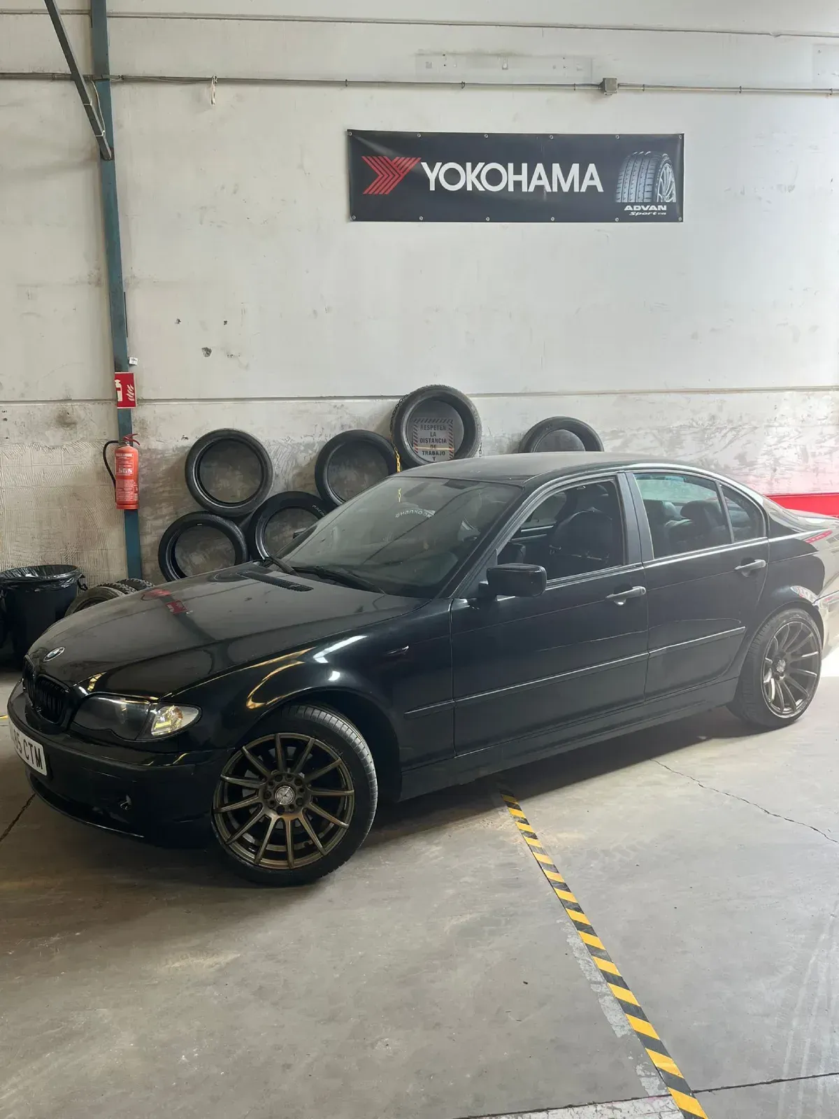 Sedán BMW negro con ruedas de bronce en un garaje, con el cartel de Yokohama al fondo.
