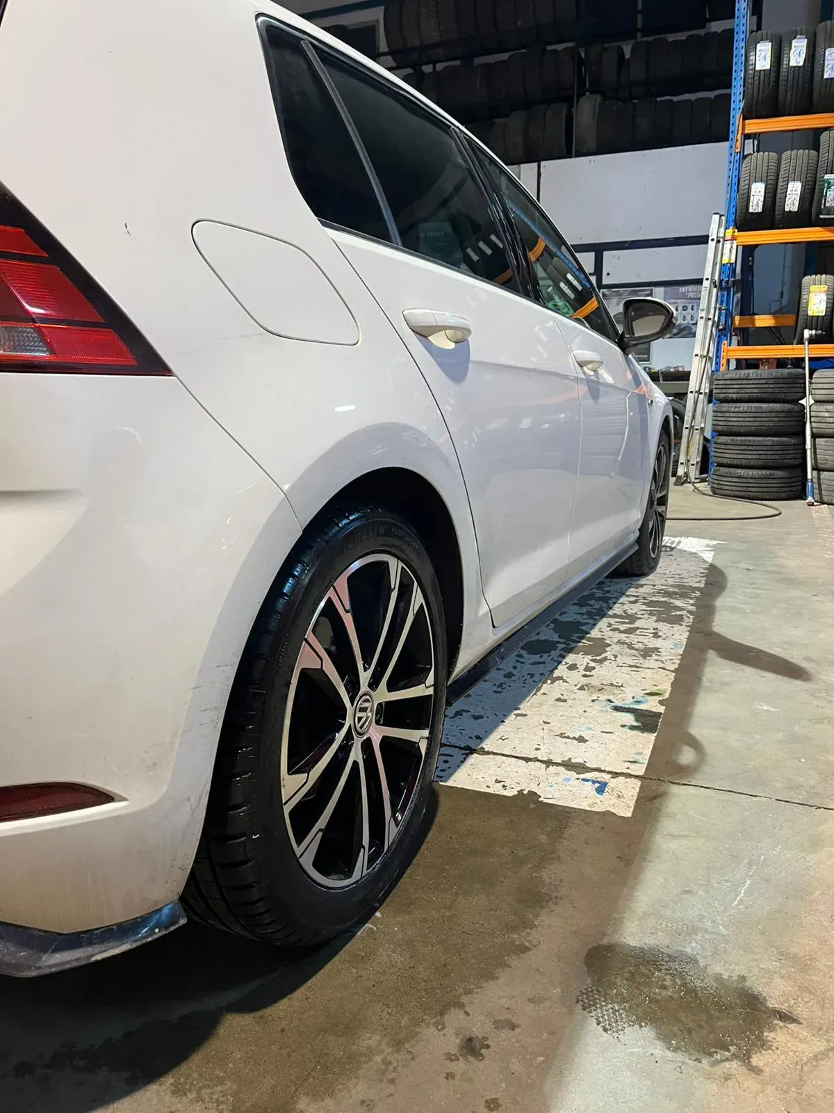 Coche blanco con llantas negras y plateadas aparcado dentro de una tienda con neumáticos apilados en el fondo.