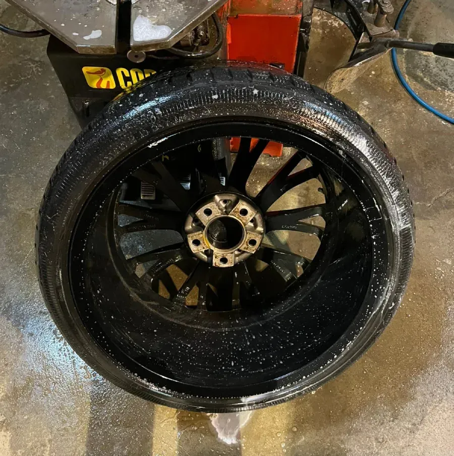 Neumático de automóvil negro con banda de rodadura sobre llanta negra, enjabonado, apoyado en el suelo de un taller.