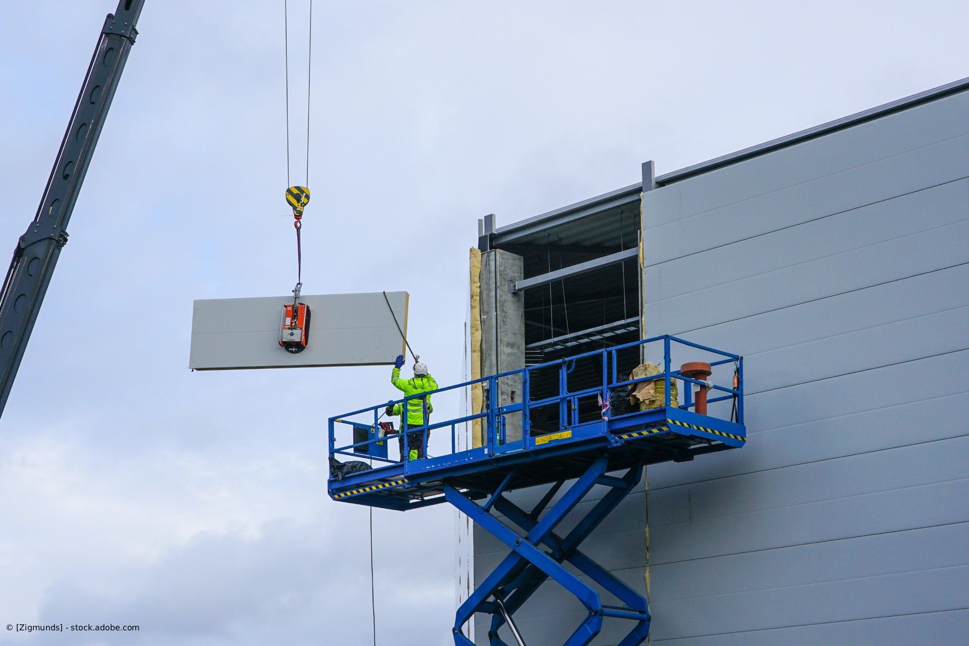 Ein Bauarbeiter auf einer Hebebühne führt eine Betonwandplatte, die von einem Kran in das Gebäude gehoben wird.