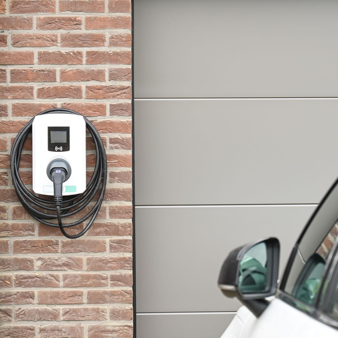 Borne de recharge électrique fixée sur un mur en briques, avec un câble enroulé, à côté d’une voiture garée devant une porte de garage.