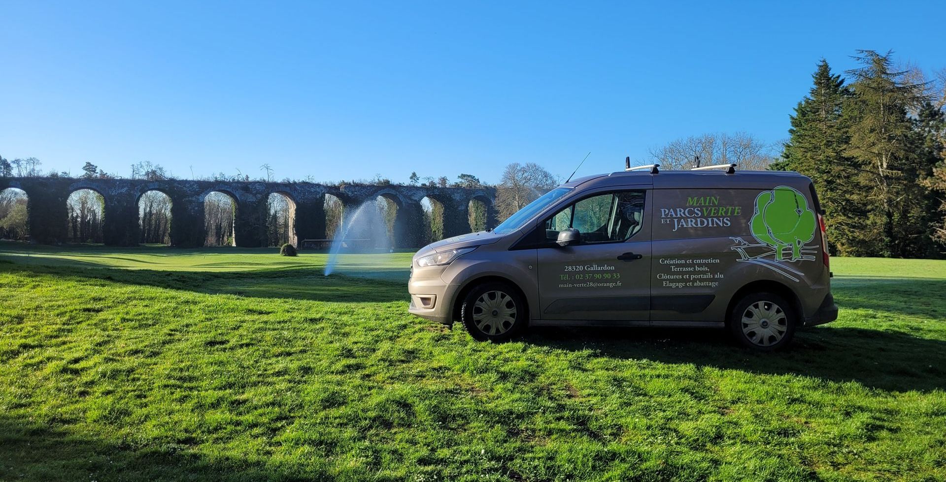 Un arboriste en pantalon et casque orange, retenu par des cordes, taille une branche d'arbre sur fond de ciel bleu.