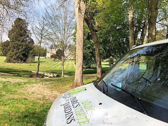 Camionnette blanche avec le logo « Parcs et Jardins » dans un parc avec des arbres et un bâtiment au loin.