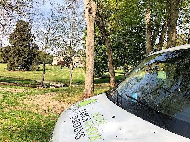 Un véhicule blanc portant l'inscription « PARCS ET JARDINS » est stationné dans un parc verdoyant avec des arbres et un bâtiment en arrière-plan.