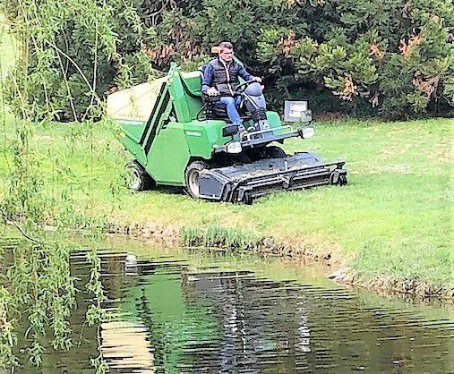 Une personne utilise une tondeuse à gazon près du bord de l'eau. La machine coupe l'herbe ; son reflet est visible dans l'eau.