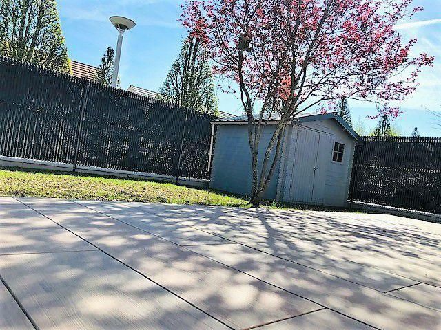 Un hangar gris à côté d'un arbre en fleurs, se détachant sur une clôture noire et un ciel dégagé.