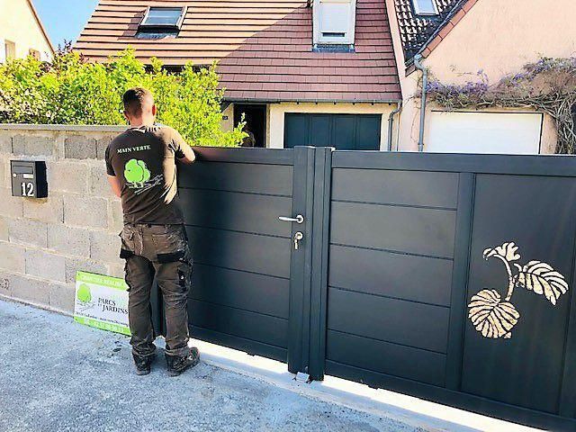 Un homme se tient près d'un portail gris foncé orné d'un autocollant représentant une plante, devant une maison.