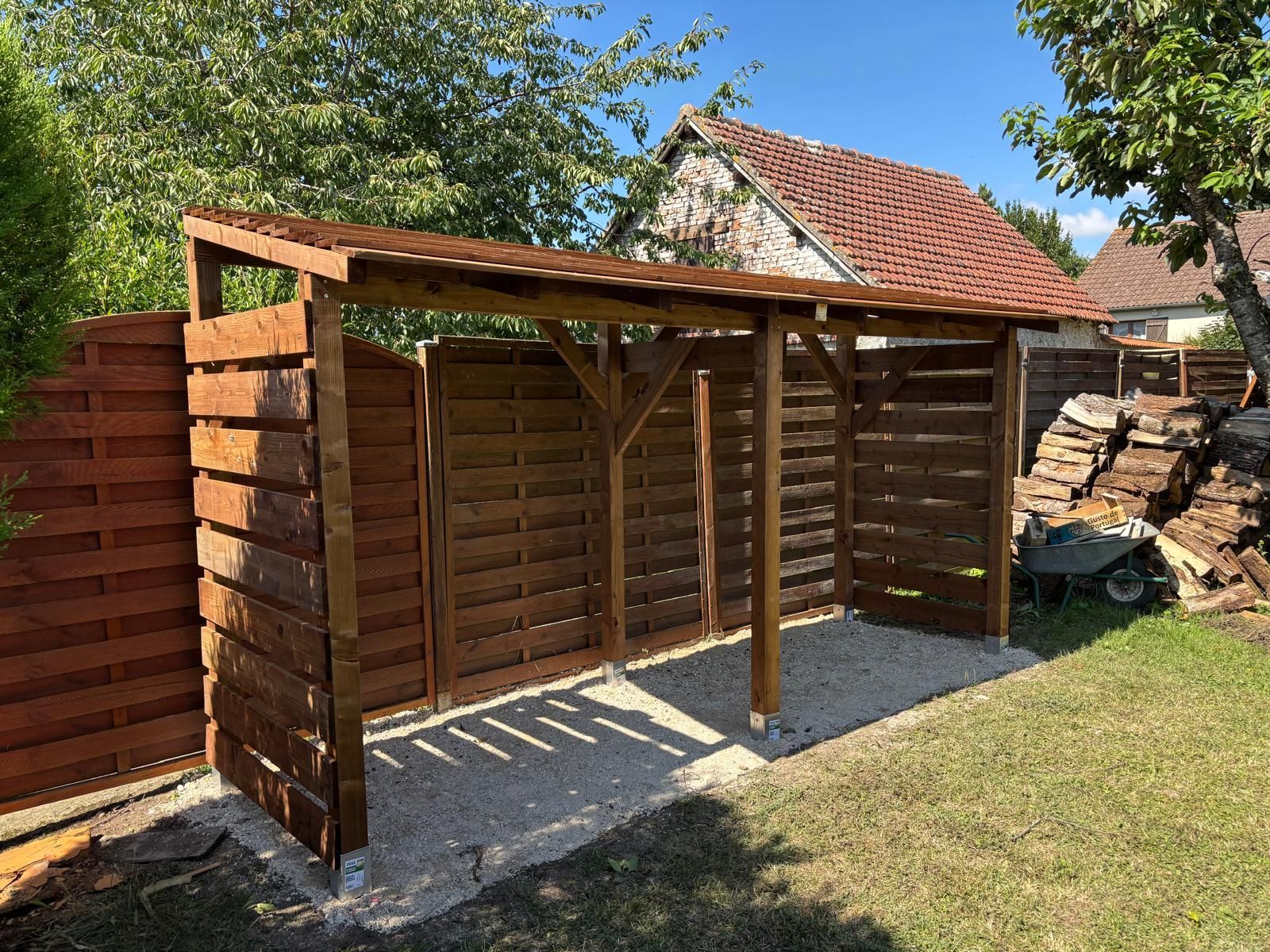 Un abri de jardin vert avec pergola se trouve dans une cour arrière, à côté d'une terrasse en béton et d'une clôture en bois, sous un ciel bleu.
