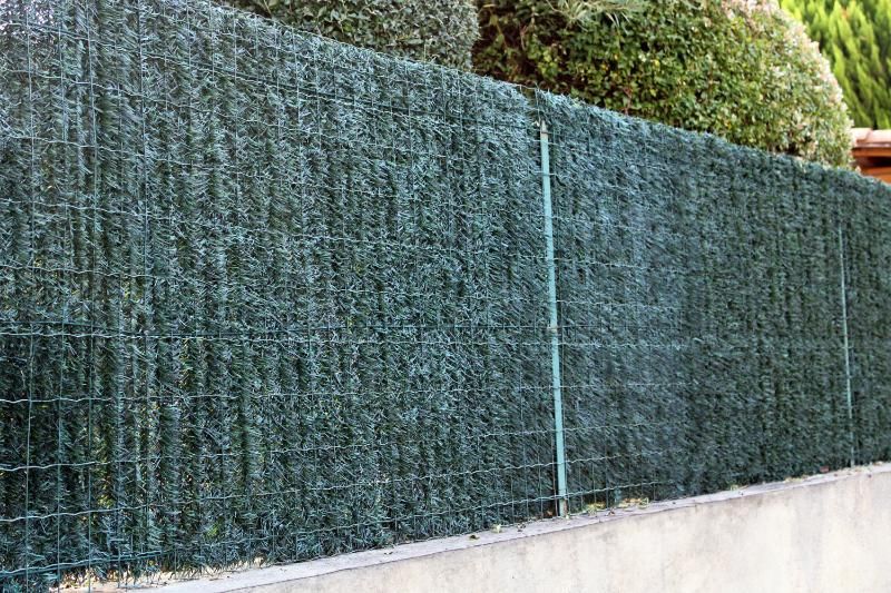 Clôture d'intimité verte composée de feuillage artificiel sur un treillis métallique, longeant un muret en béton.