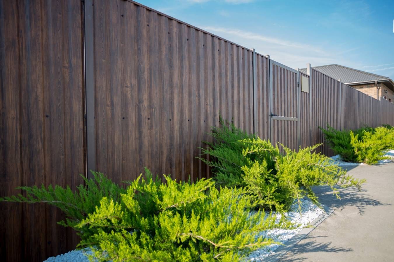 Clôture en tôle ondulée marron avec des buissons verts le long d'un chemin de pierres blanches.