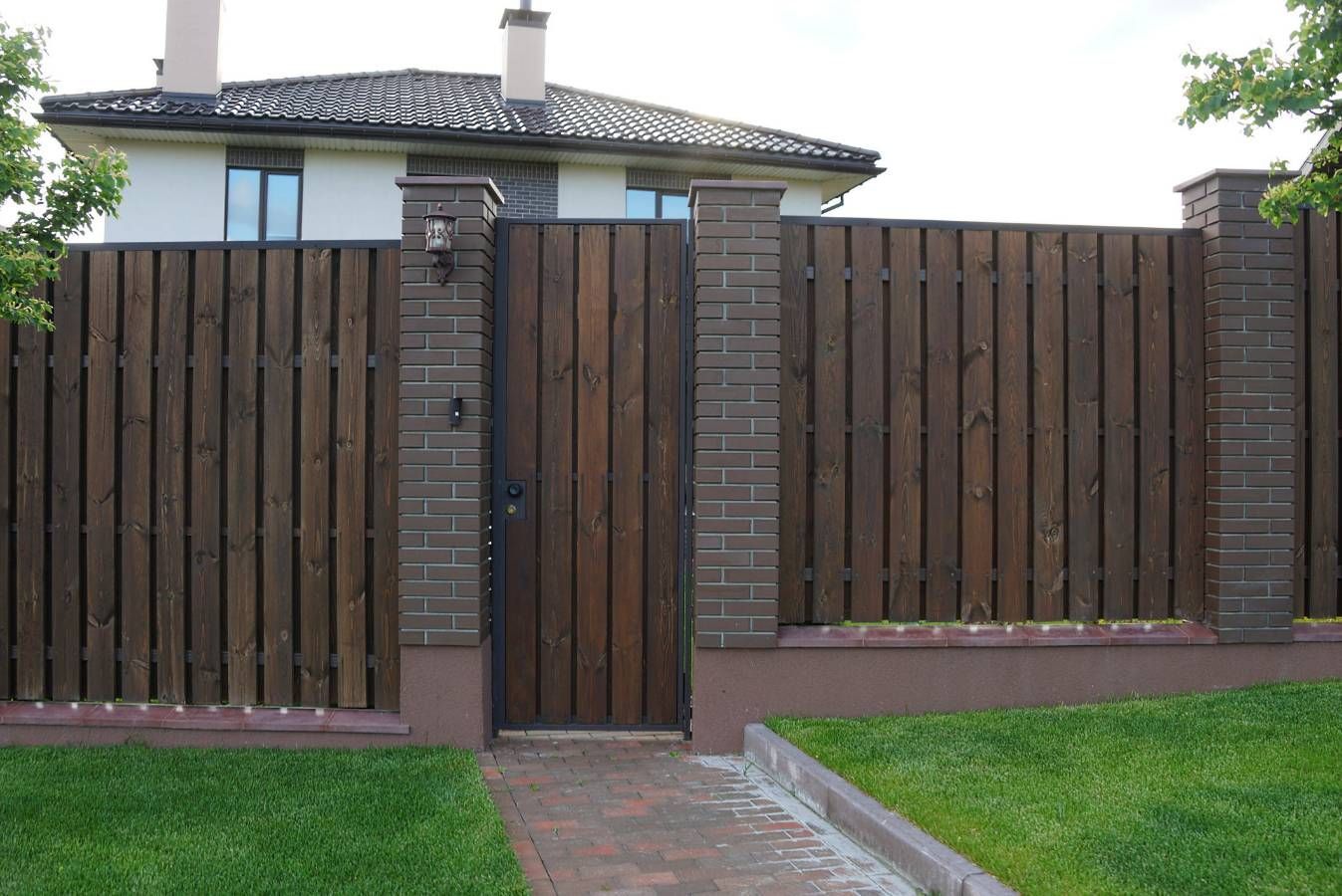 Clôture en bois avec piliers en briques et portail devant une maison, allée en briques et pelouse verte.