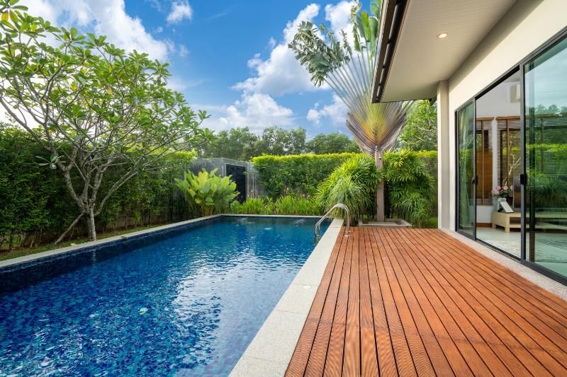 Piscine jouxtant une terrasse en bois et une maison aux portes vitrées ; végétation luxuriante et ciel bleu.