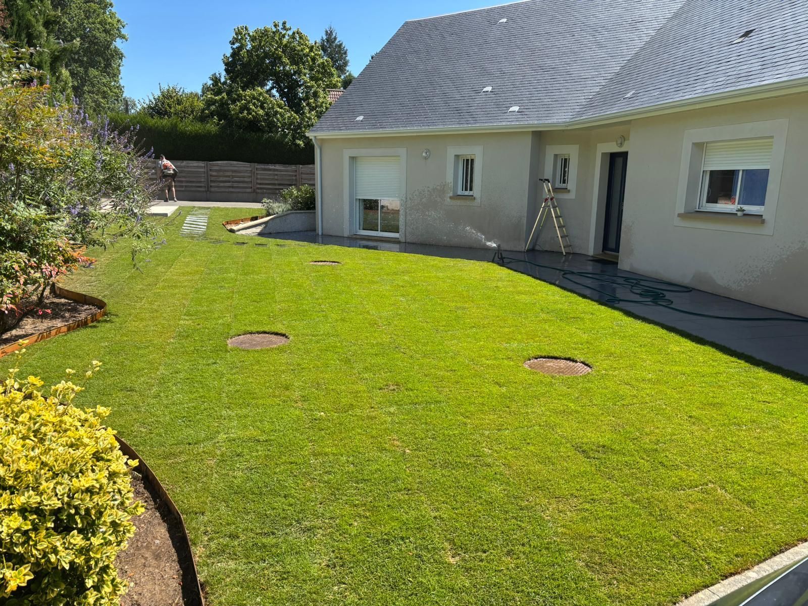 Pelouse verte dans un jardin, avec une maison en arrière-plan. Quelqu'un arrose la pelouse.