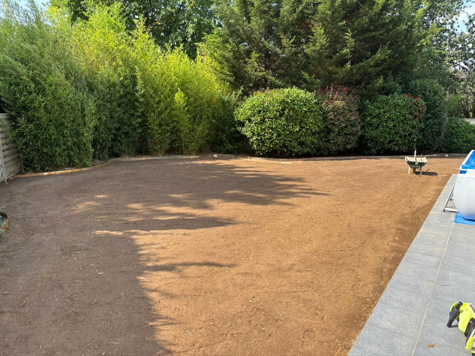 Une cour dégagée à la terre brune, entourée de buissons verts et d'une terrasse en pierre.