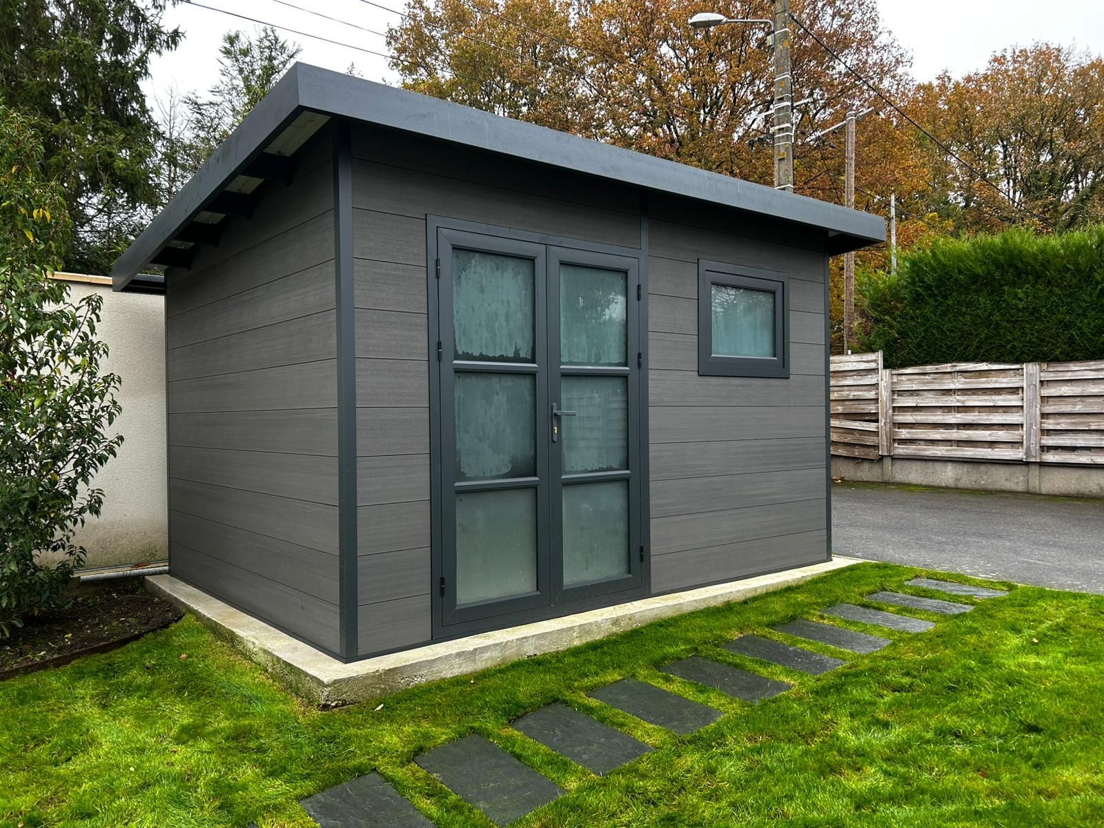 Abri de jardin en bois avec murs et toit à claire-voie, sur un sol gravillonné, adjacent à une clôture en bois, à l'extérieur.