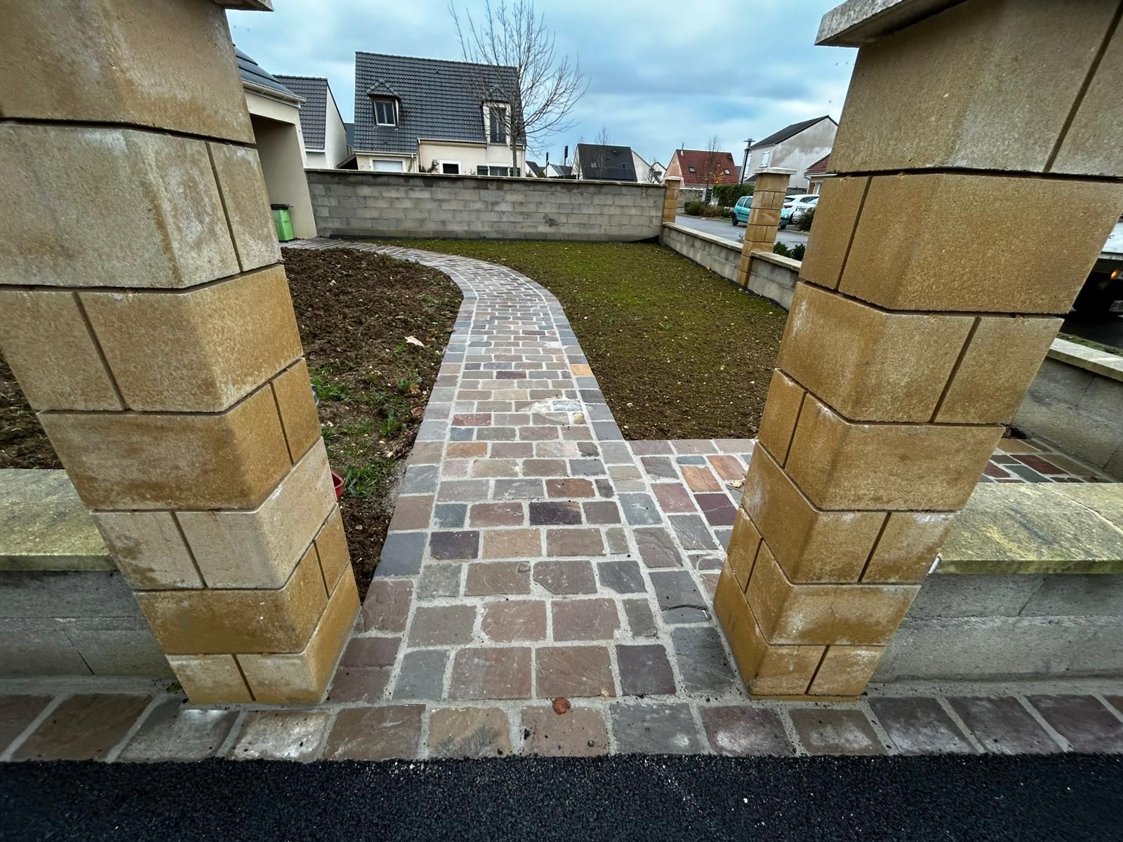 Allée bordée de pierres décoratives et de plantes, menant à une rue où se trouve une voiture.