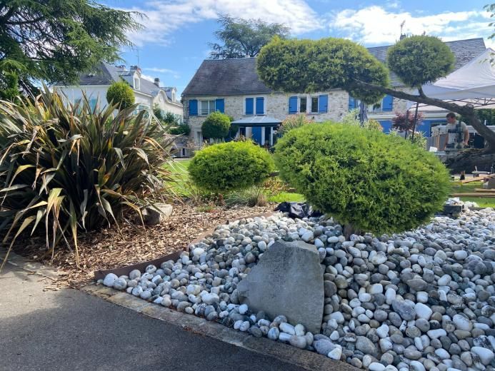 Cour avant aménagée avec une maison en pierre en arrière-plan. Arbustes verts et pierres blanches dans le jardin.