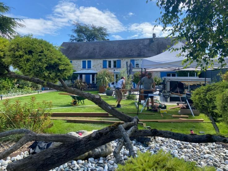 Deux ouvriers travaillent dans un jardin, une maison se dresse en arrière-plan. Pelouse verte, volets bleus, journée ensoleillée.