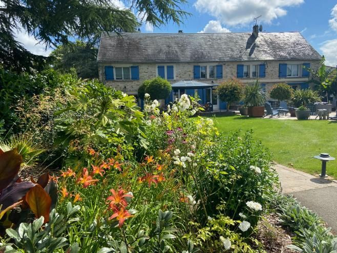 Maison en pierre aux volets bleus, entourée d'un jardin luxuriant aux feuillages orange, blancs et verts, par une journée ensoleillée.