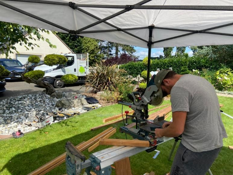 Un homme coupe du bois à la scie sous un auvent, à l'extérieur, avec une pelouse et un jardin verts en arrière-plan.