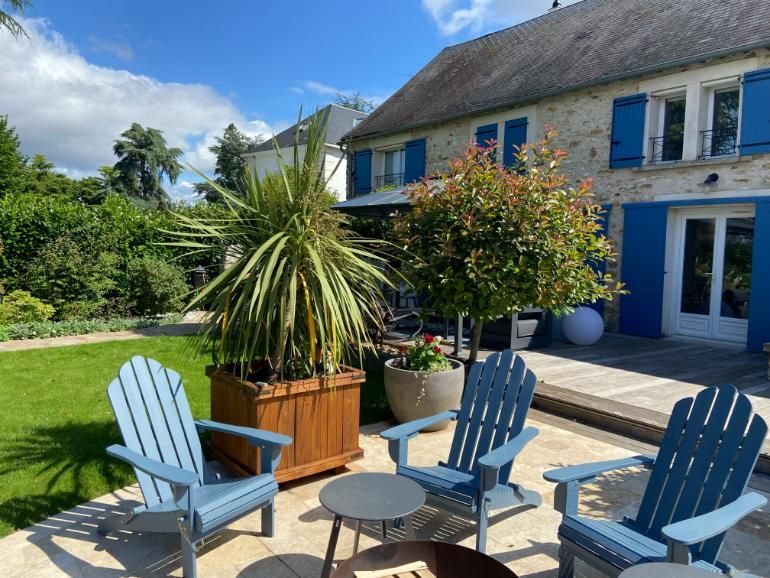 Trois chaises Adirondack bleues sur une terrasse, face à une maison aux volets bleus. Pelouse verte et arbres.