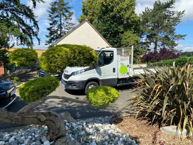 Un camion blanc est garé dans une allée, avec des arbres et des buissons verts devant une maison, sous un ciel bleu.