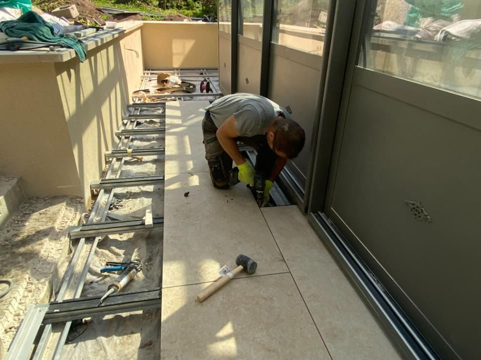 Un ouvrier du bâtiment pose du carrelage dans une véranda. Il utilise des outils et porte des gants.