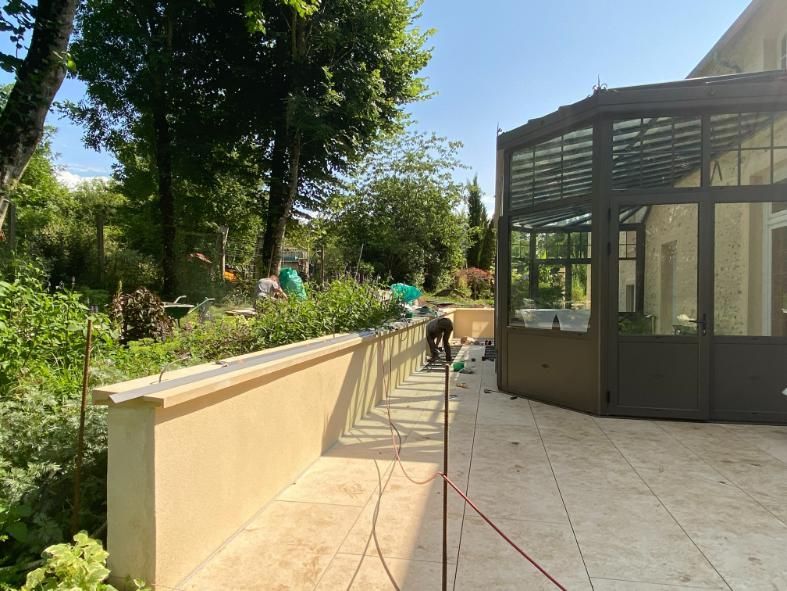 Jardin avec terrasse, pelouse et maison. Table et chaises sur la terrasse. Journée ensoleillée.