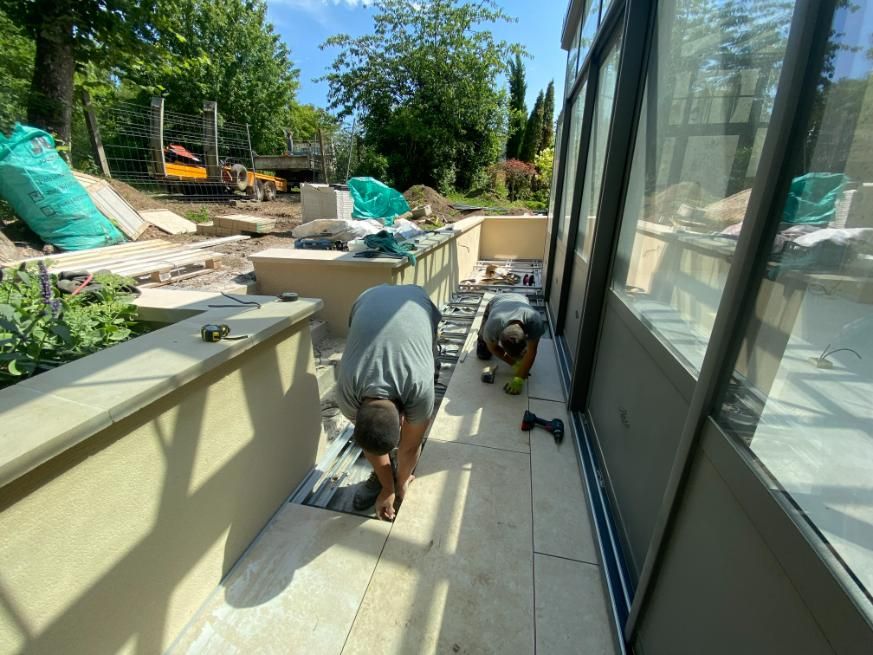 Deux ouvriers posent du carrelage sur une terrasse jouxtant une paroi vitrée. On aperçoit des matériaux et des outils de construction.