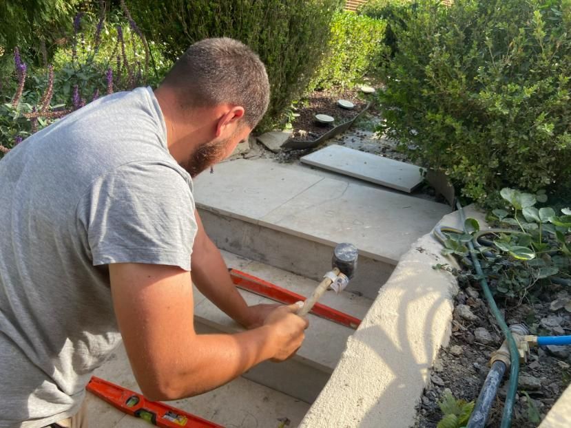 Un homme utilise un marteau pour travailler sur des marches en pierre à l'extérieur, avec un niveau et des buissons.