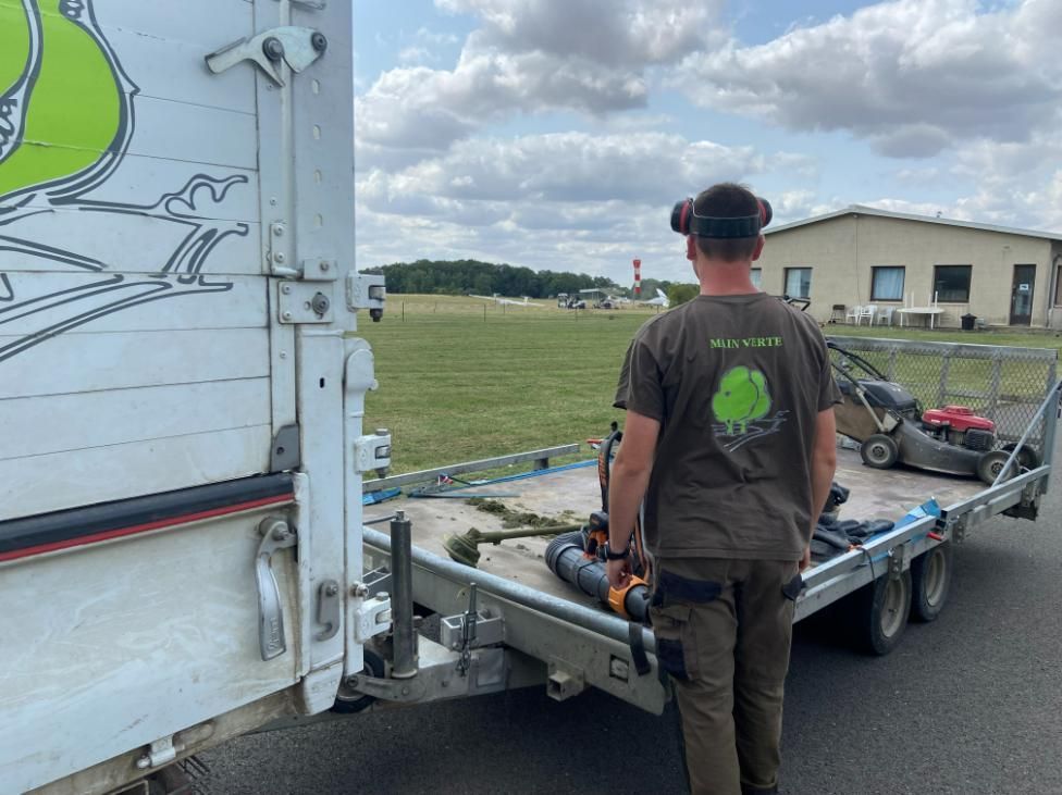 Un arboriste en pantalon et harnais orange élague une branche d'arbre sur fond de ciel bleu.